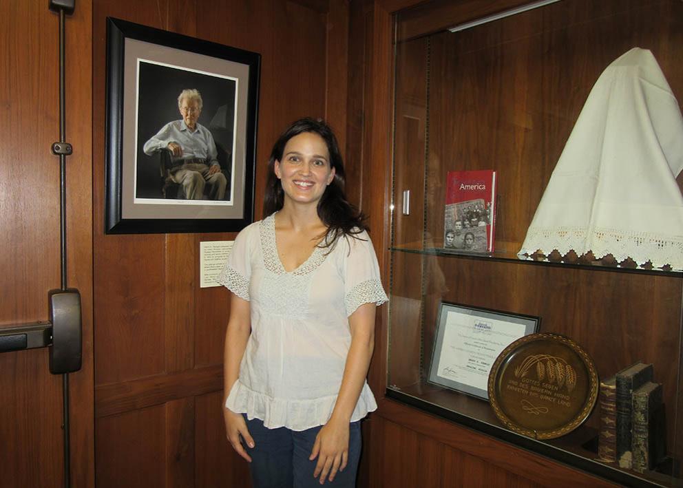woman in white shirt standing in front of wood paneled wall with a framed photo on her right and gallery display on her left